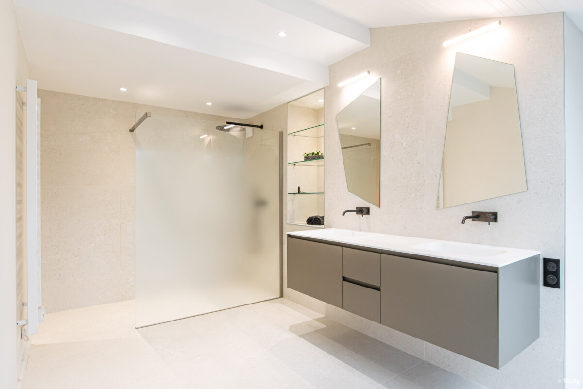 Idée déco salle de bain nature, salle de bain minimaliste épurée, douche italienne paroi verre opaque dépoli, pommeau encastré inox, meuble double vasque suspendu gris taupe mat, plan de travail blanc, double miroir polygonal asymétrique, robinet mural noir mat, carrelage pierre naturelle beige travertin, éclairage LED encastré, sol béton ciré blanc
