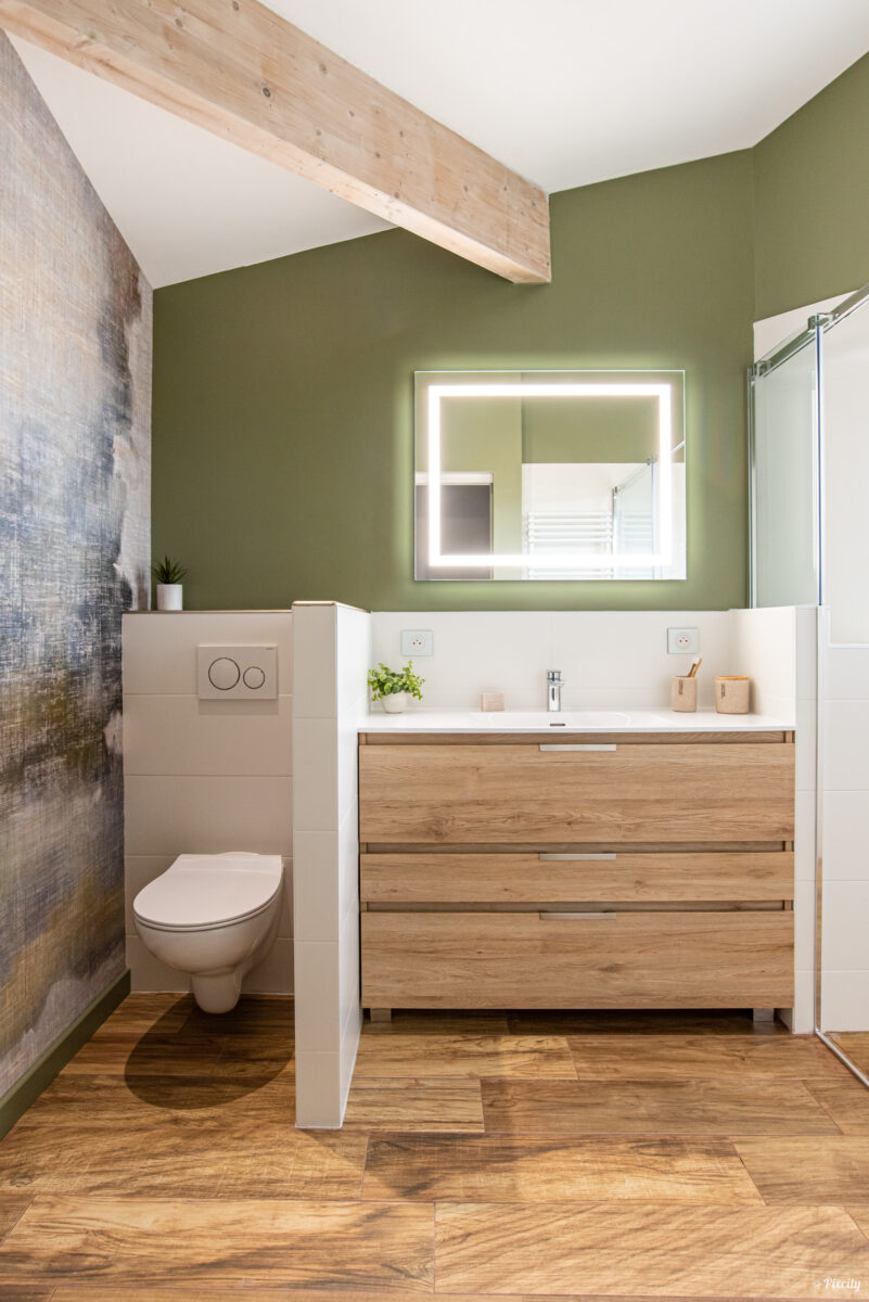 Idée déco salle de bain nature sous pente, meuble vasque bois chêne naturel tiroirs, plan de travail blanc, miroir LED rectangulaire rétroéclairé, WC suspendu blanc, poutre bois brut apparent, mur vert olive, papier peint panoramique paysage abstrait naturel, sol parquet bois chevrons, plante verte pot blanc, accessoires rose poudré
