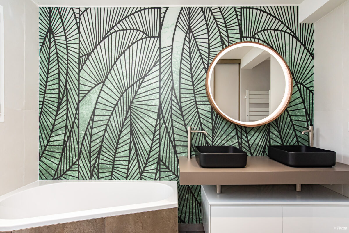 Idée déco salle de bain nature, papier peint panoramique feuilles tropicales vert et noir, double vasque noire rectangulaire à poser, meuble suspendu blanc tiroirs, plan de travail beige taupe, miroir rond rétroéclairé LED encadrement bois cuivré, baignoire encastrée blanche, robinetterie inox brossé