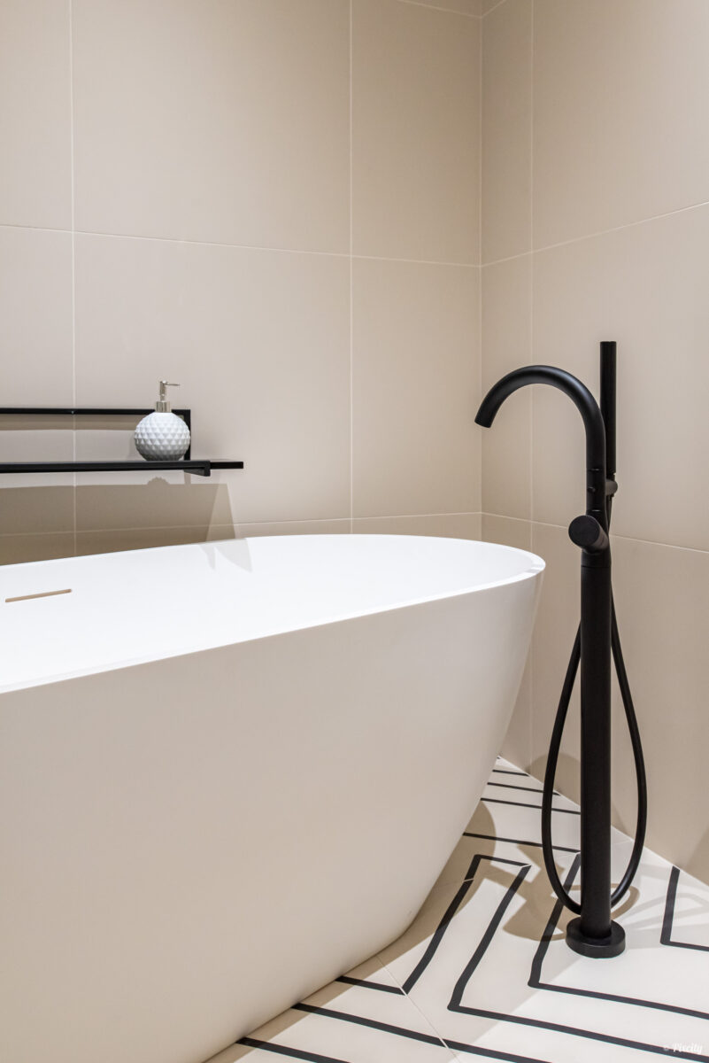 Idée déco salle de bain nature, détail baignoire îlot ovale blanche, robinet sur pied colonne métal noir mat, tablette murale étagère métal noir, distributeur savon céramique blanc, sol carrelage géométrique noir et blanc graphique, carrelage mural beige grand format