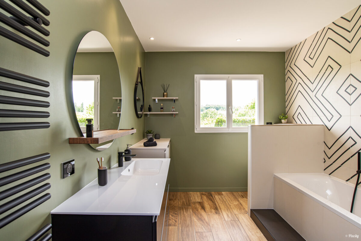 Idée déco salle de bain nature, vue d'ensemble, baignoire encastrée blanche, douche italienne paroi verre, radiateur sèche-serviettes noir mat, meuble double vasque blanc, miroir ovale LED rétroéclairé, étagères bois, mur vert sauge, sol parquet bois naturel, fenêtres lumière naturelle, carrelage géométrique noir et blanc