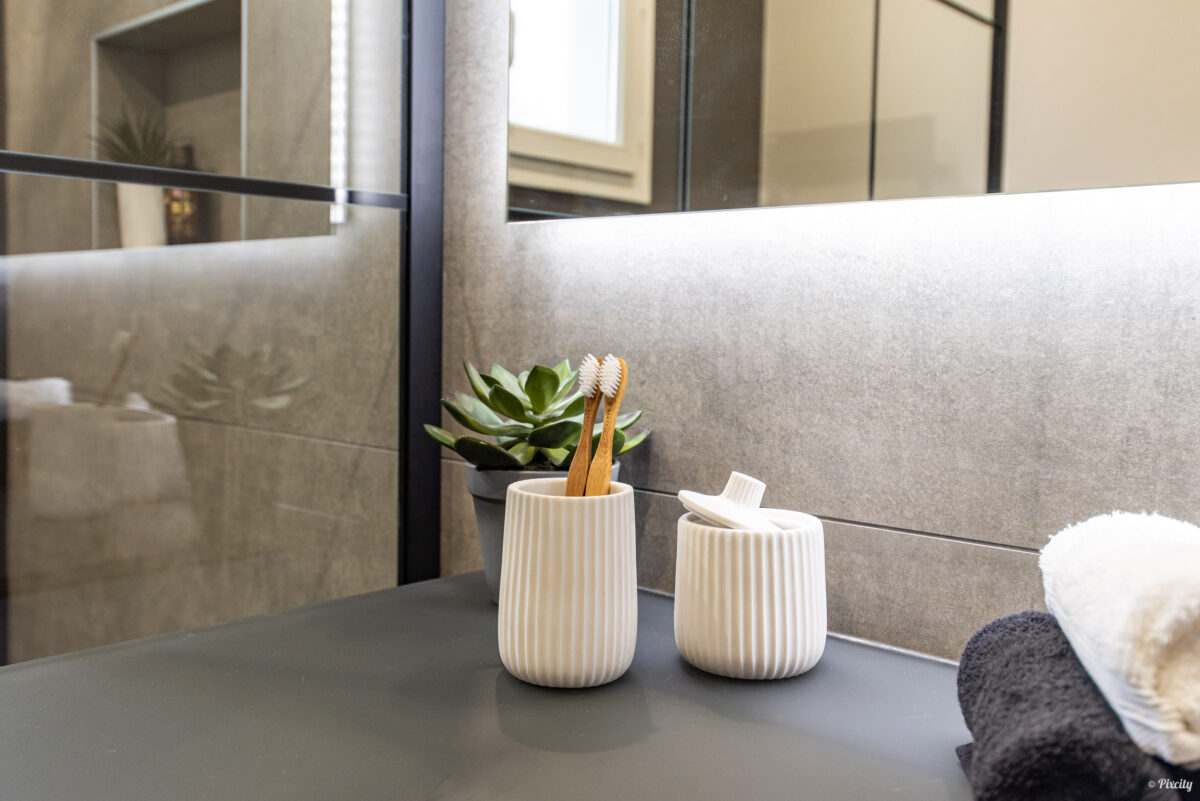 Idée déco salle de bain nature, détail accessoires, pot à brosses bambou blanc cannelé avec brosses bambou écologiques, pot accessoire blanc, plante succulente verte, serviettes anthracite et blanches moelleuses, plan de travail gris foncé mat, miroir rétroéclairé, carrelage gris pierre, armoire miroir étagères