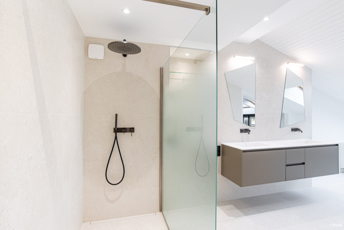 Idée déco salle de bain nature, douche italienne pommeau encastré rond noir mat, carrelage pierre naturelle beige travertin, paroi verre transparent, meuble double vasque suspendu gris taupe, plan de travail blanc, double miroir forme originale, robinet mural noir, éclairage LED encastré, sol béton ciré