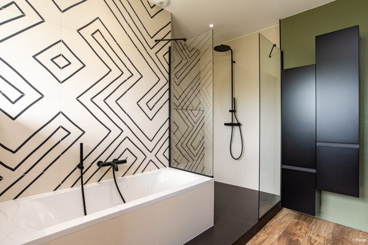 Idée déco salle de bain nature, détail douche italienne, carrelage géométrique graphique noir et blanc, pommeau colonne noir mat, paroi verre transparent, meuble colonne noir mat, sol imitation parquet bois, mur vert olive, robinetterie noire mate