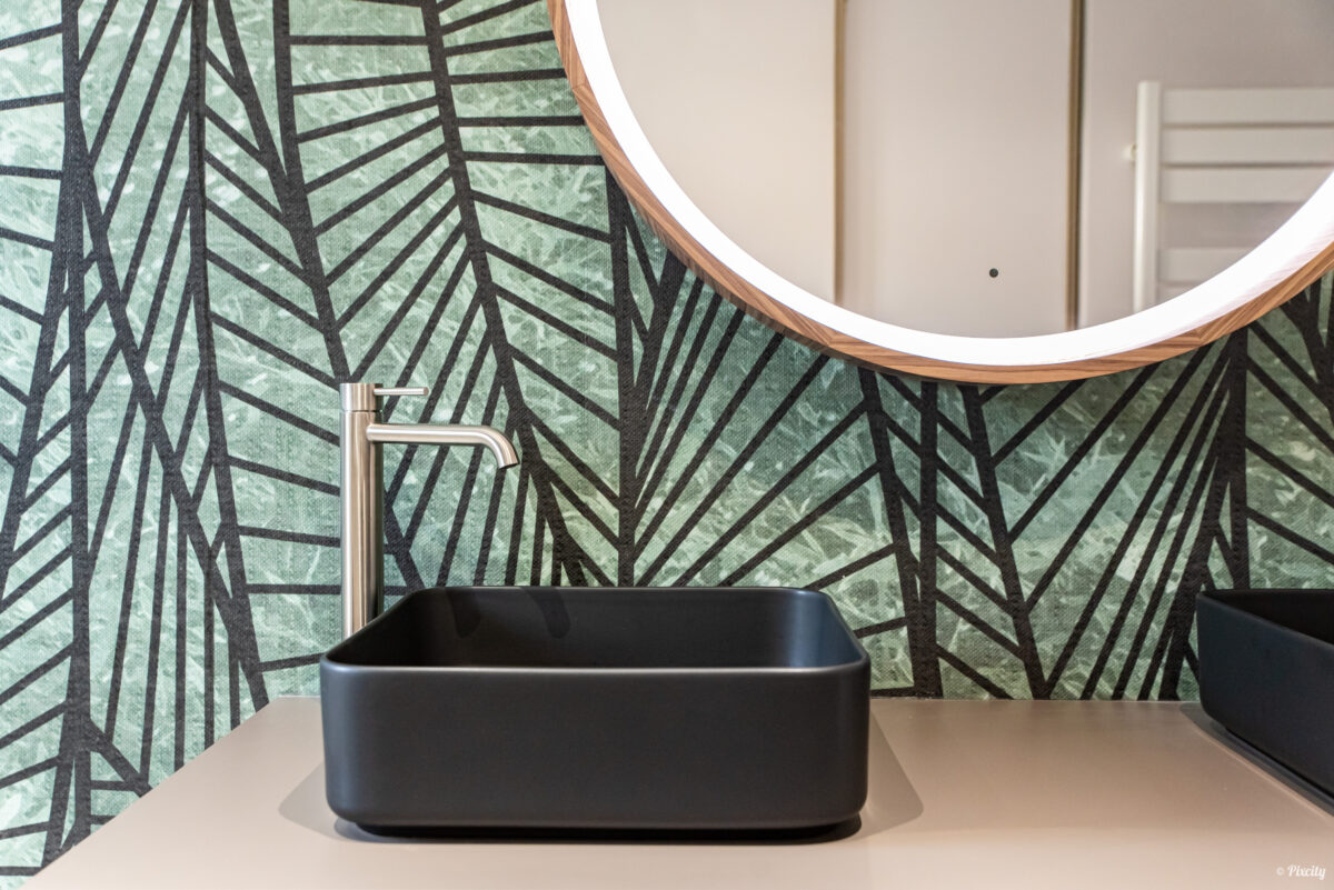 Idée déco salle de bain nature, détail vasque rectangulaire noire à poser sur plan de travail beige, robinet haut mitigeur inox brossé, papier peint feuilles tropicales vert et noir, miroir rond rétroéclairé cadre bois cuivré, meuble suspendu blanc laqué