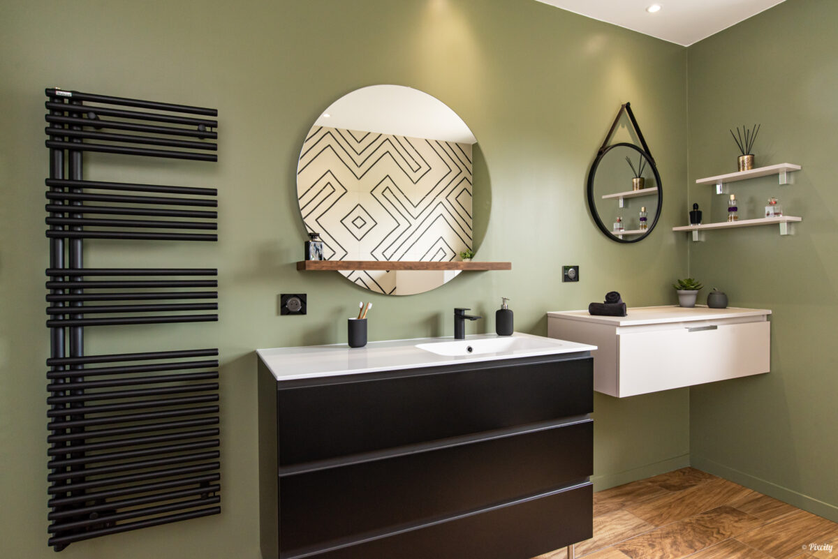 Idée déco salle de bain nature, vue panoramique, mur vert kaki olive, meuble double vasque noir mat, double miroir rond, radiateur sèche-serviettes noir mat vertical, plan de travail bois naturel, étagères murales blanches parfums plantes, sol parquet bois effet naturel, carrelage géométrique graphique noir et blanc