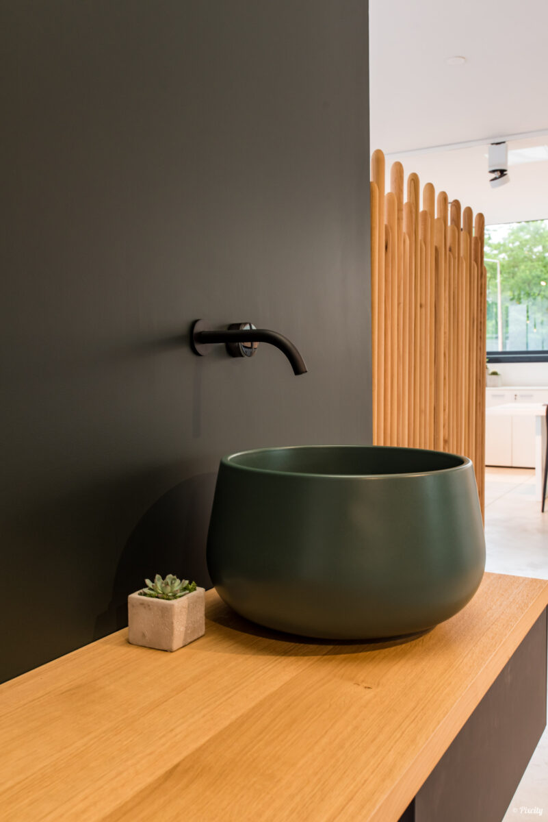 Idée déco salle de bain nature, vasque ronde bol céramique vert kaki mat, plan de travail bois chêne clair massif, robinet mural noir mat, cloison séparatrice noire, claustra lattes bois naturel, plante succulente pot béton, lumière naturelle fenêtre