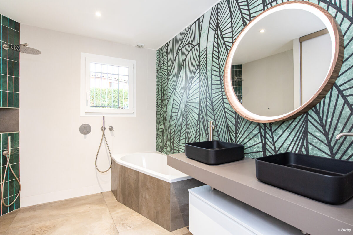 Idée déco salle de bain nature, papier peint tropical feuilles de bananier vert et noir grand format, double vasque rectangulaire noire à poser, meuble suspendu blanc laqué, plan de travail béton gris, miroir rond rétroéclairé LED encadrement bois cuivré, baignoire encastrée blanche, carrelage zellige vert émeraude colonne, sol pierre naturelle beige, robinetterie inox brossé, fenêtre lumière naturelle
