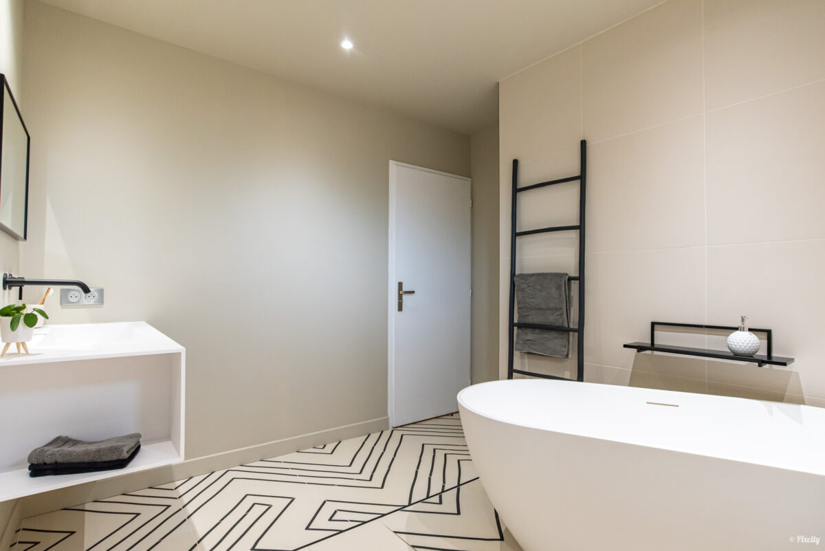 Idée déco salle de bain nature, salle de bain épurée scandinave, baignoire îlot ovale blanche, porte-serviettes échelle métal noir mat, meuble vasque blanc cube ouvert, robinet mural noir mat, serviette grise, miroir carré cadre noir, plante verte pot blanc, sol carrelage chevron géométrique noir et blanc graphique