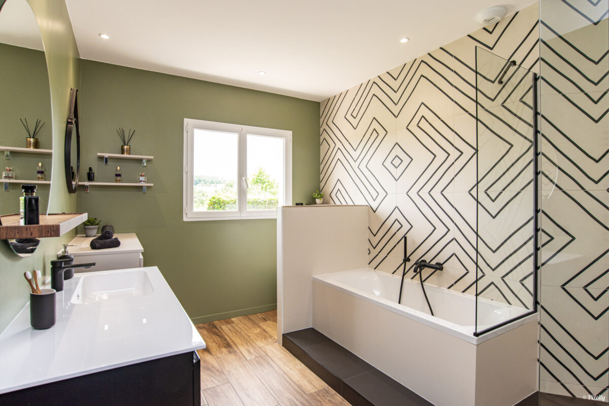 Idée déco salle de bain nature, mur vert kaki olive, carrelage mural géométrique graphique noir et blanc, baignoire encastrée blanche avec pare-baignoire verre, double vasque encastrée plan de travail blanc, meuble suspendu noir mat, étagères murales bois naturel, flacons parfum diffuseurs, plantes vertes, sol imitation parquet bois, fenêtre lumière naturelle