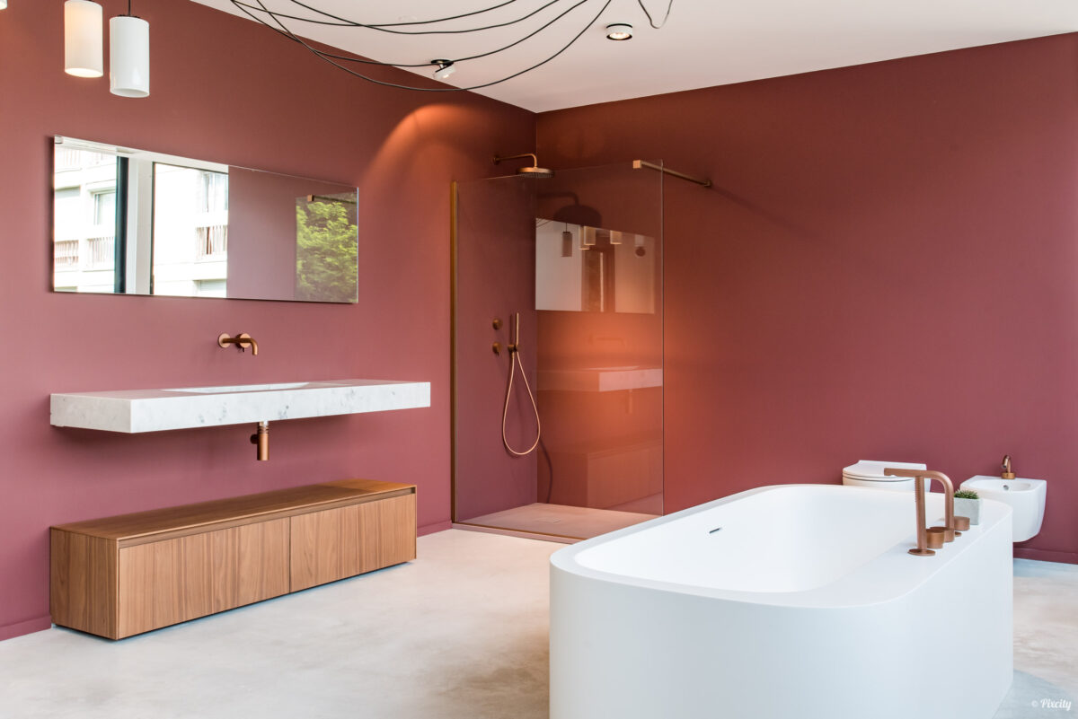 Idée déco salle de bain nature, murs rouge bordeaux terracotta, baignoire îlot ovale blanche sur béton ciré, vasque marbre blanc rectangulaire suspendue, meuble bas bois naturel chêne, robinetterie dorée laiton, douche italienne paroi verre fumé, tête de douche cuivre, suspension luminaire blanc, fenêtre lumière naturelle