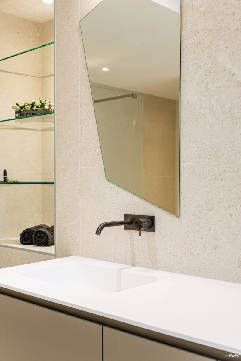 Idée déco salle de bain nature, détail vasque encastrée blanche, miroir asymétrique polygonal, robinet mural noir brossé, étagères verre transparent encastrées, plantes vertes en pot, serviettes noires roulées, carrelage pierre naturelle beige travertin