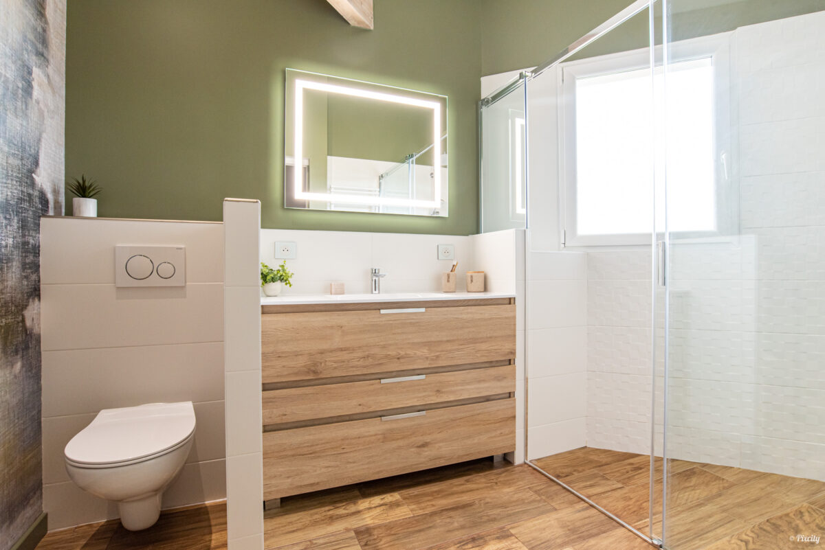Idée déco salle de bain nature sous pente vue d'ensemble, meuble vasque bois chêne naturel, plan de travail blanc, miroir LED rétroéclairé, WC suspendu, douche italienne paroi verre coulissante, mur vert olive, papier peint panoramique paysage abstrait, sol parquet bois chevrons, fenêtre lumière naturelle, plante verte