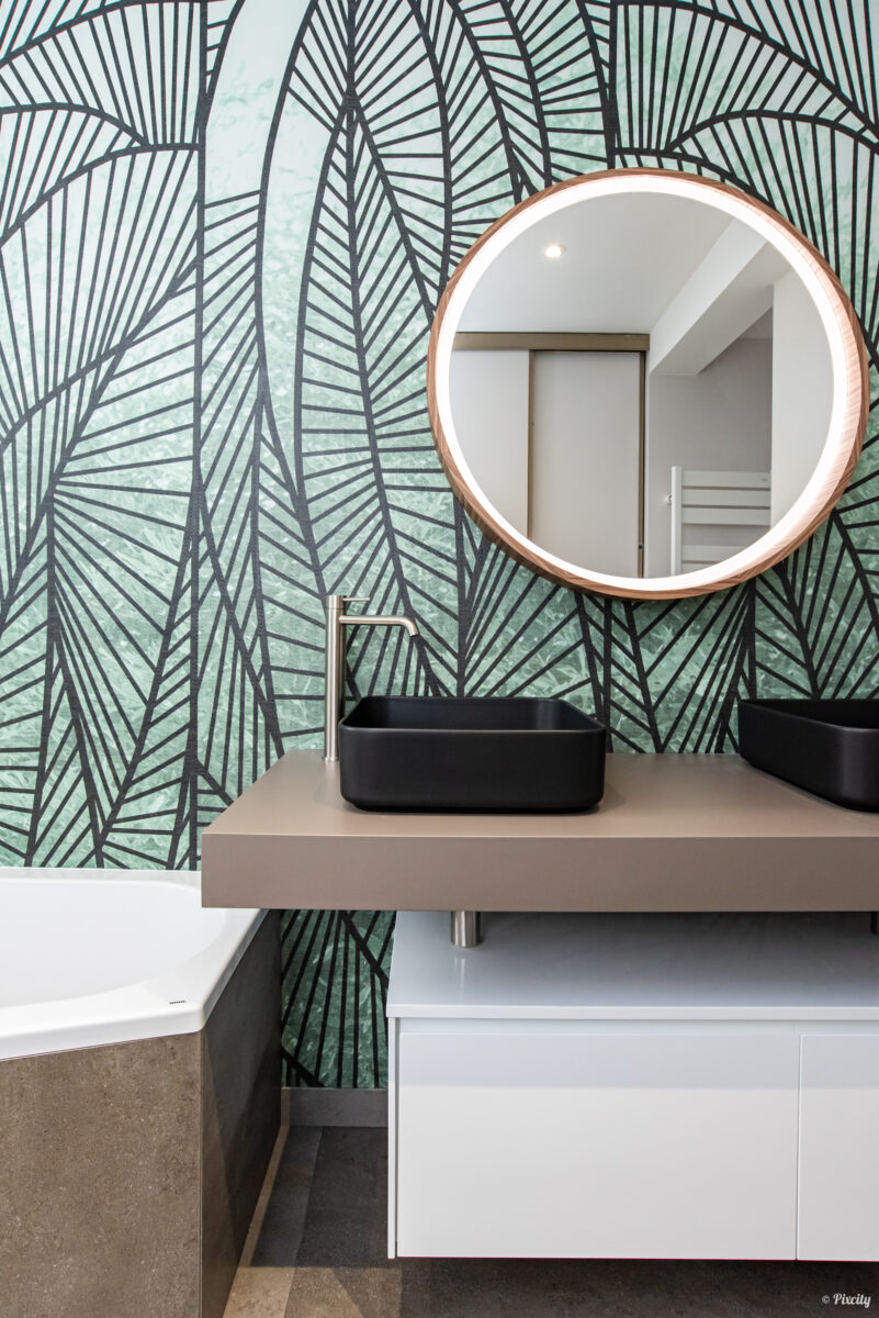 Idée déco salle de bain nature, papier peint feuilles de palmier vert et noir, vasque rectangulaire noire à poser, robinet mitigeur haut inox brossé, meuble suspendu blanc laqué, plan de travail beige taupe, miroir rond rétroéclairé LED cadre bois cuivré, baignoire blanche
