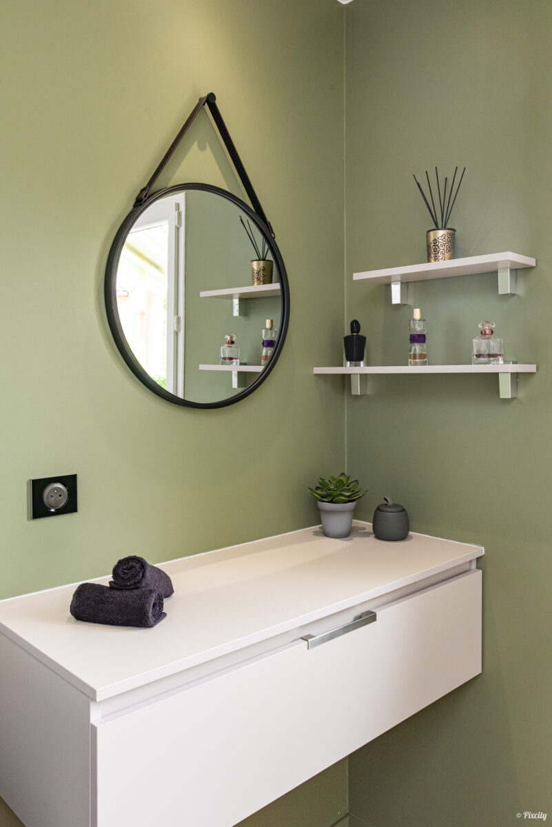 Idée déco salle de bain nature, détail mur vert sauge olive, miroir rond style Adnet sangle cuir noir, étagères murales blanches, diffuseur parfum bâtons dorés, flacons parfum colorés, succulente pot gris, bougie noire, serviette anthracite pliée, meuble vasque blanc mat