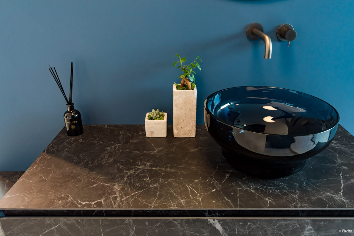 Idée déco salle de bain nature, détail plan de travail marbre noir veiné, vasque ronde en verre bleu nuit brillant, robinet mural bronze brossé, diffuseur parfum bâtons noirs, plante succulente en pot béton, bonsaï en pot travertin beige, mur bleu canard