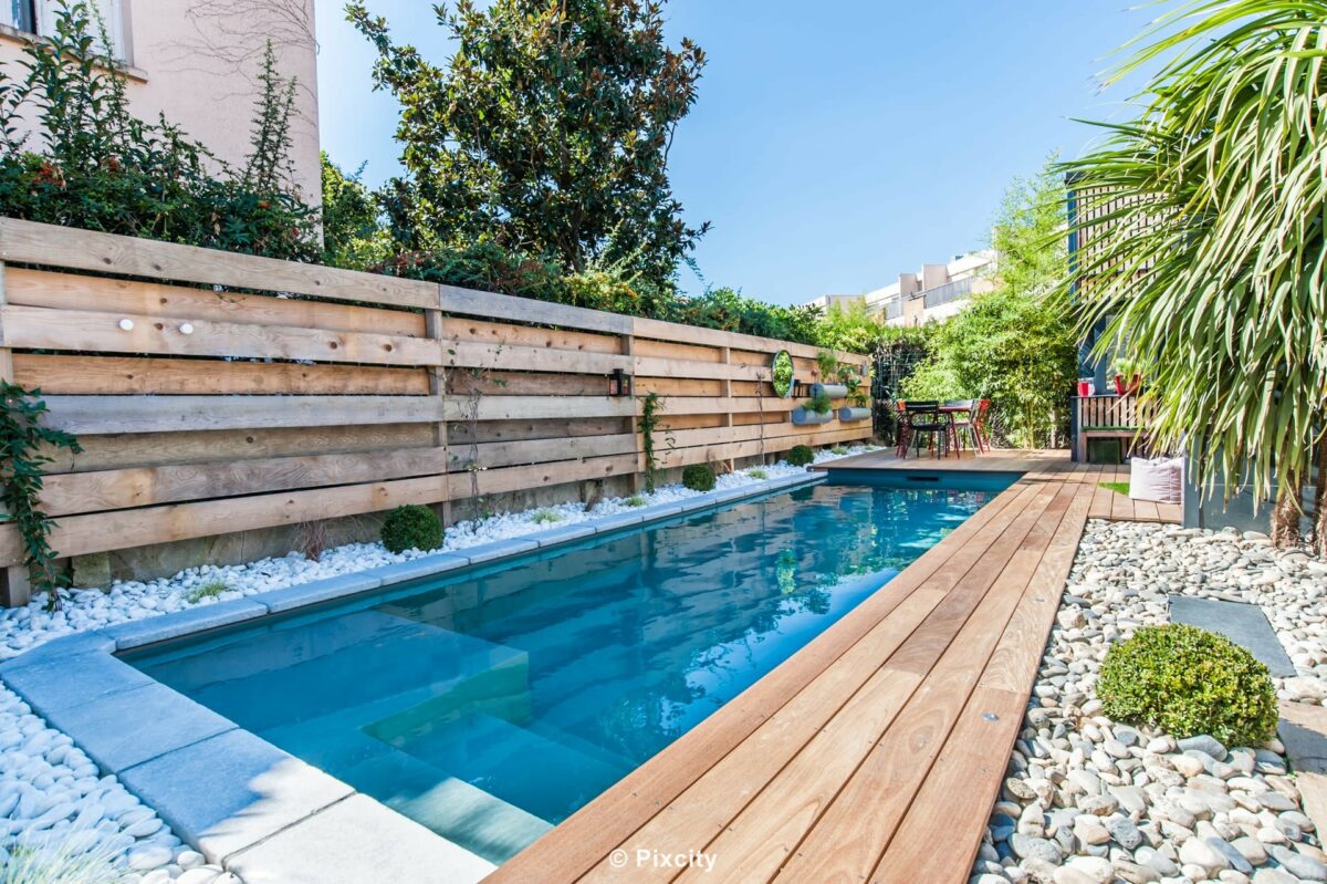 Aménagement terrasse bois autour piscine, piscine couloir rectangulaire eau bleu ardoise, terrasse bois exotique brun chaud, palissade bois horizontal clair, galets blancs décoratifs, palmier trachycarpus, buis en boule, pergola bois, style méditerranéen contemporain, ciel bleu estival