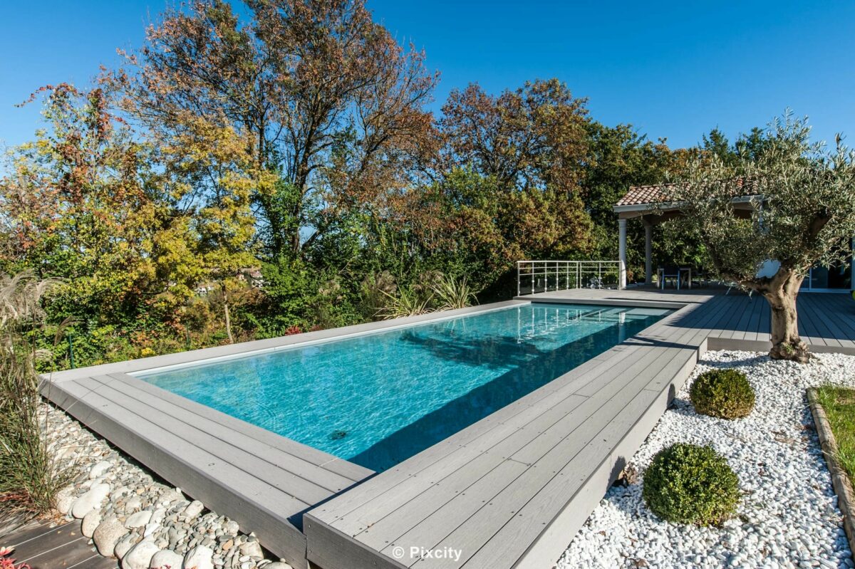 Aménagement terrasse bois autour piscine, piscine rectangulaire eau bleu turquoise, terrasse composite gris clair, arbres feuillage automne orange et rouge, buis sphériques taillés, olivier adulte, pergola bois tonnelle, galets blancs, ciel bleu, ambiance automne sud de France