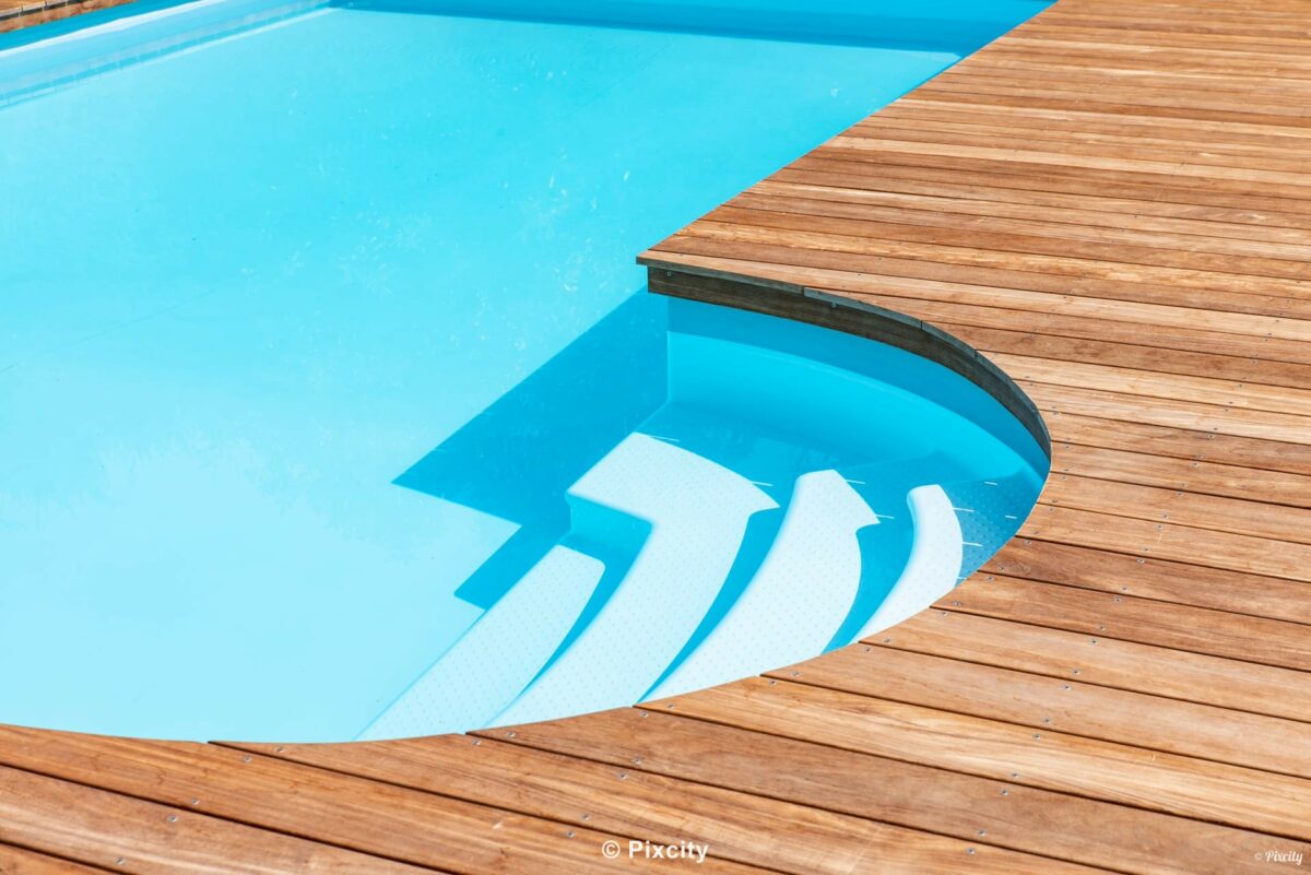 Aménagement terrasse bois autour piscine, gros plan escalier roman immergé marches courbes blanches piscine, terrasse bois exotique doré, eau bleu turquoise translucide, reflets ombres géométriques, détail architecture piscine, jeux de lumière rasante
