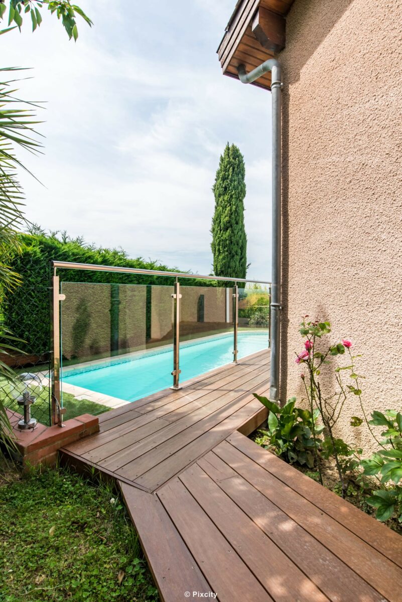 Aménagement terrasse bois autour piscine, terrasse bois exotique brun clair, garde-corps inox et verre trempé, piscine rectangulaire eau bleu-vert, cyprès méditerranéen, rosier grimpant fleurs roses, mur enduit beige, palmier trachycarpus, pelouse, ciel nuageux