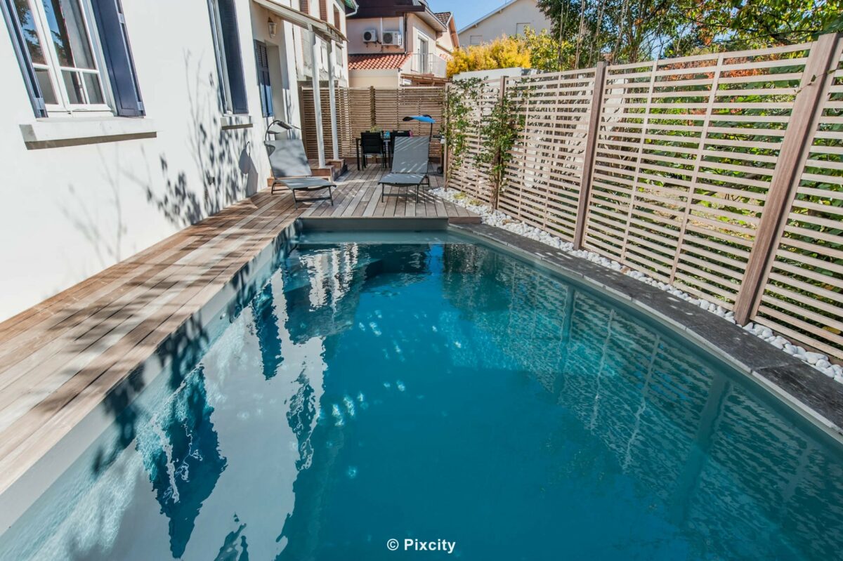 Aménagement terrasse bois autour piscine, piscine rectangulaire étroite eau bleu foncé reflets, terrasse composite gris brun, chaises longues design noires, brise-vue aluminium lattes grises, maison contemporaine urbaine, jardin citadin intimiste, ciel bleu été