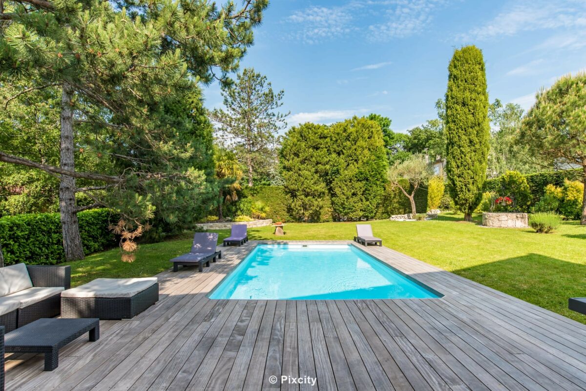 Aménagement terrasse bois autour piscine, grande piscine rectangulaire eau cristalline turquoise, terrasse bois exotique clair très spacieuse, transats noirs design, salon jardin gris anthracite, cyprès topiaires, pins sylvestres, pelouse verte irriguée, ciel bleu, ambiance mas provençal luxueux