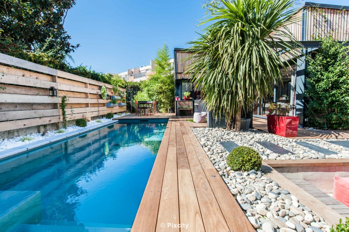 Aménagement terrasse bois autour piscine, piscine couloir longue eau bleu sombre reflets, terrasse bois exotique chaleureux, galets blancs décoratifs, palmier trachycarpus, plante en pot rouge vif, pergola bois poteaux, brise-vue lattes horizontales bois, végétation tropicale, ciel bleu vif