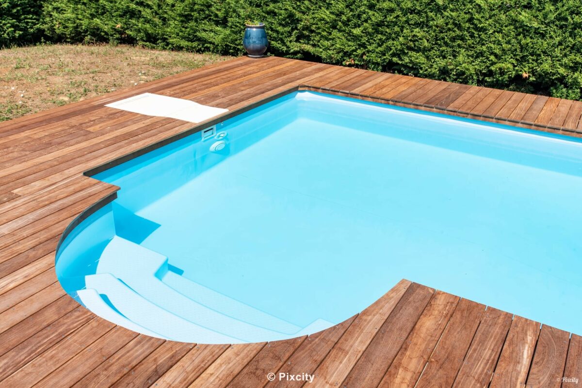 Aménagement terrasse bois autour piscine, piscine rectangulaire arrondie eau bleue, terrasse bois teck brun doré, haie verte épaisse taillée, pelouse verte, pot céramique bleu artisanal, bords nets, lumière douce été, ambiance jardin champêtre