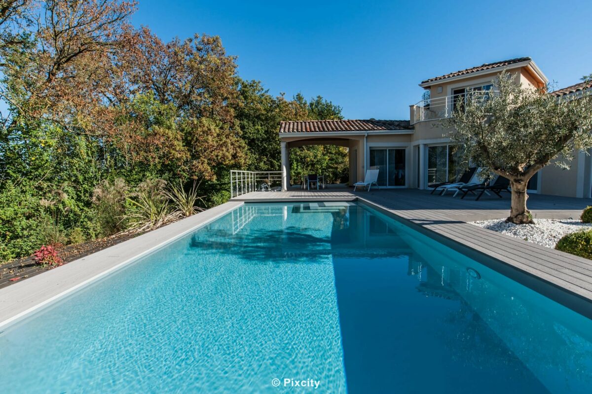 Aménagement terrasse bois autour piscine, vue large villa provençale toits tuiles, piscine rectangulaire eau bleue, terrasse composite gris anthracite, palmier trachycarpus, grand pin parasol, chaises longues bois, pelouse, maison résidentielle, ciel bleu, automne