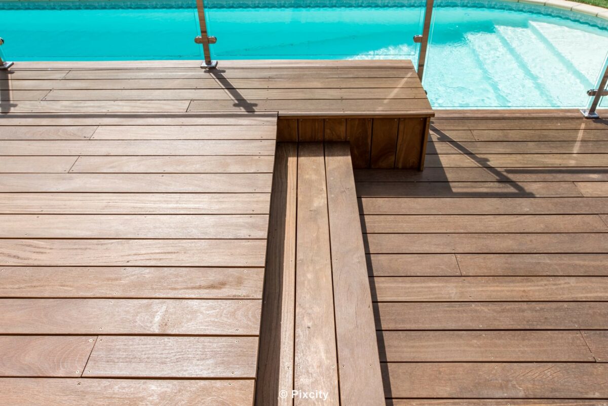 Aménagement terrasse bois autour piscine, gros plan terrasse bois ipé exotique brun chaud, banquette intégrée bois, garde-corps inox et verre trempé transparent, piscine eau turquoise, ombres portées graphiques, texture bois horizontal, détail menuiserie qualité, lumière directe