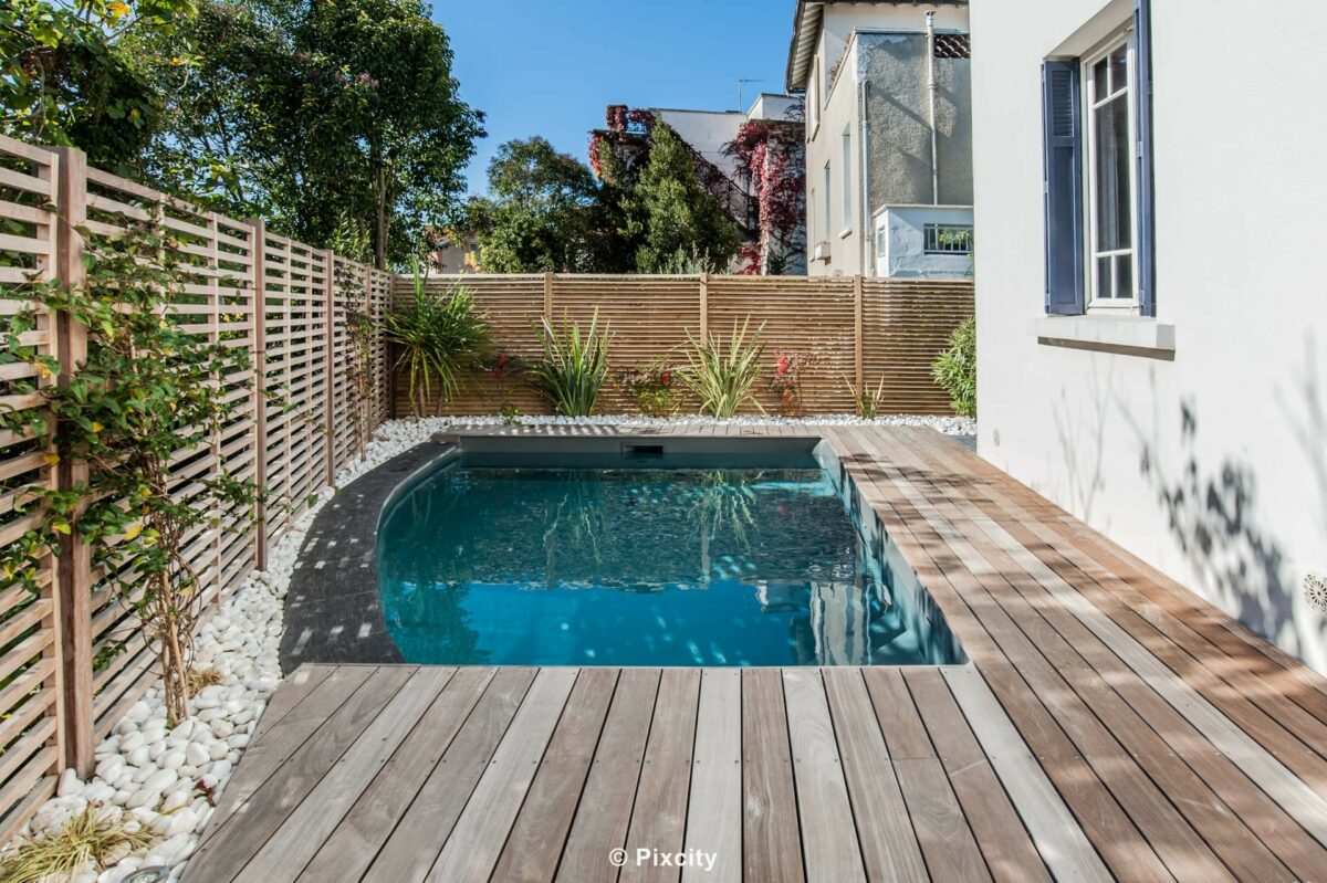 Aménagement terrasse bois autour piscine, petite piscine ovale fond gris ardoise eau bleu profond, terrasse bois gris naturel patiné, palissade bois horizontal, dracaena pot, palmier en pot, géranium rouge vif, maison blanche, jardin urbain intimiste, ciel bleu