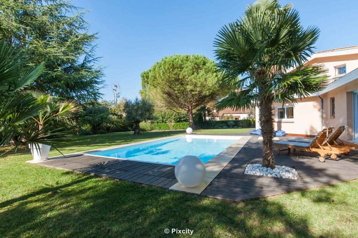 Aménagement terrasse bois autour piscine, villa provençale toits tuiles rondes, palmier trachycarpus tronc épais, grand pin parasol, piscine rectangulaire eau bleue, terrasse composite gris anthracite, chaises longues bois teck coussins, pot blanc design, pelouse verte, ciel bleu
