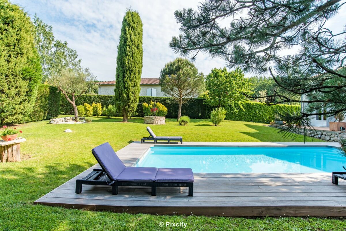 Aménagement terrasse bois autour piscine, piscine rectangulaire eau turquoise, pelouse verte impeccable, chaise longue tissu gris anthracite, cyprès méditerranéens, pins, buis taillé, terrasse bois exotique, maison résidentielle grise, ciel bleu été