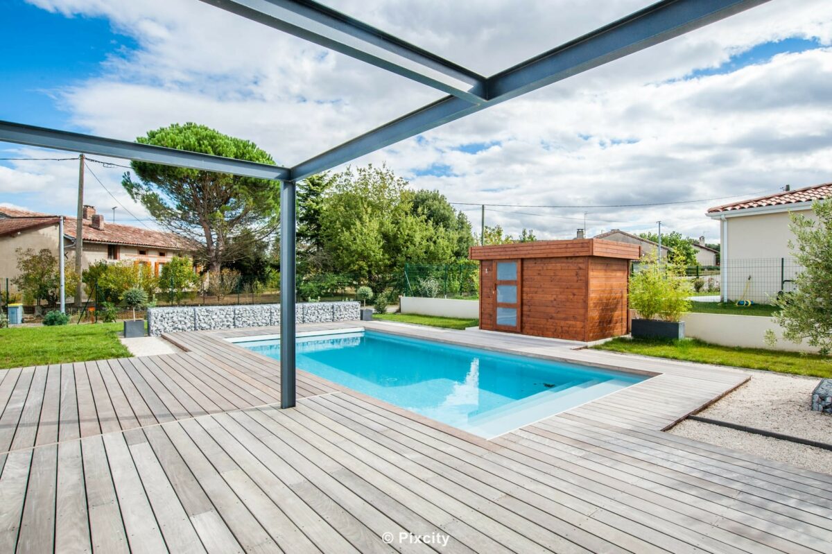 Aménagement terrasse bois autour piscine, pergola bioclimatique aluminium gris anthracite, piscine rectangulaire eau bleue, terrasse composite gris clair, abri jardin cubique bois rouge, fontaine jet d'eau piscine, pelouse verte, arbres, maison pavillon, ciel nuageux