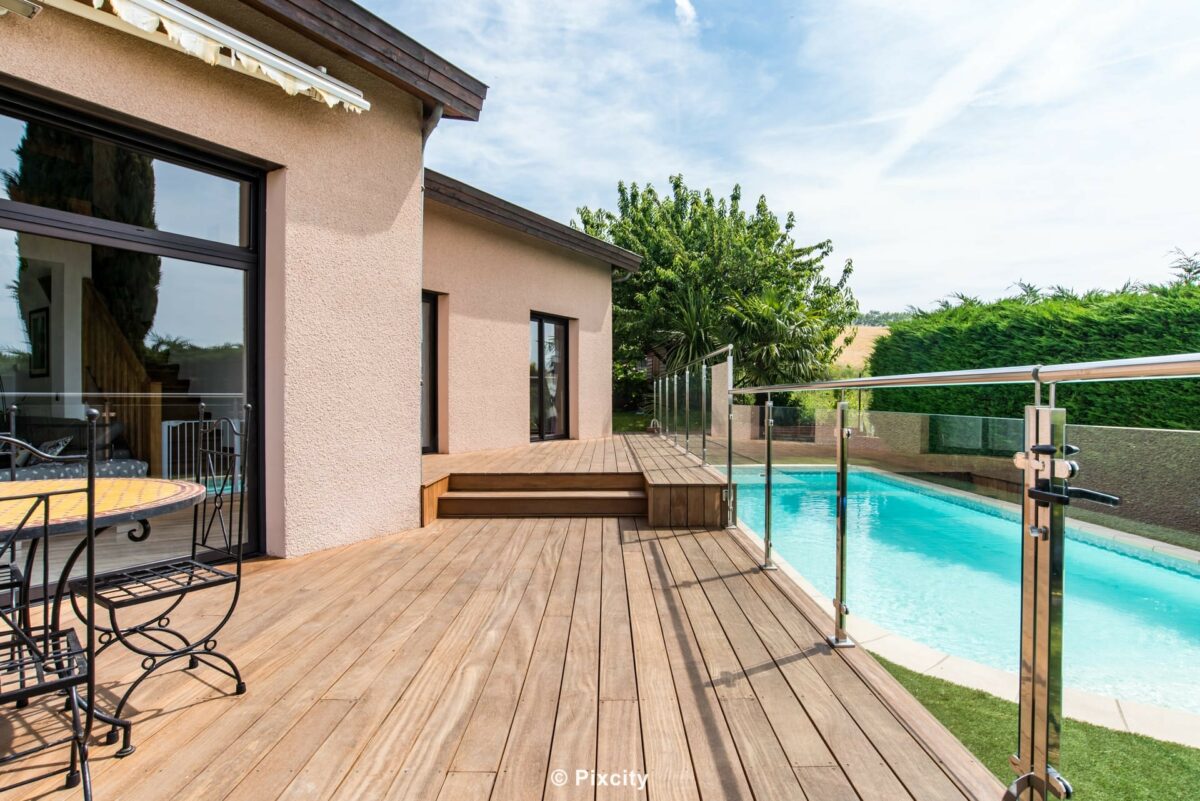 Aménagement terrasse bois autour piscine, terrasse bois exotique escalier deux niveaux, garde-corps inox et verre poli, piscine bleue en fond, maison enduit beige fenêtres aluminium noires, rosier, palmier, bougainvillier, style contemporain sudiste