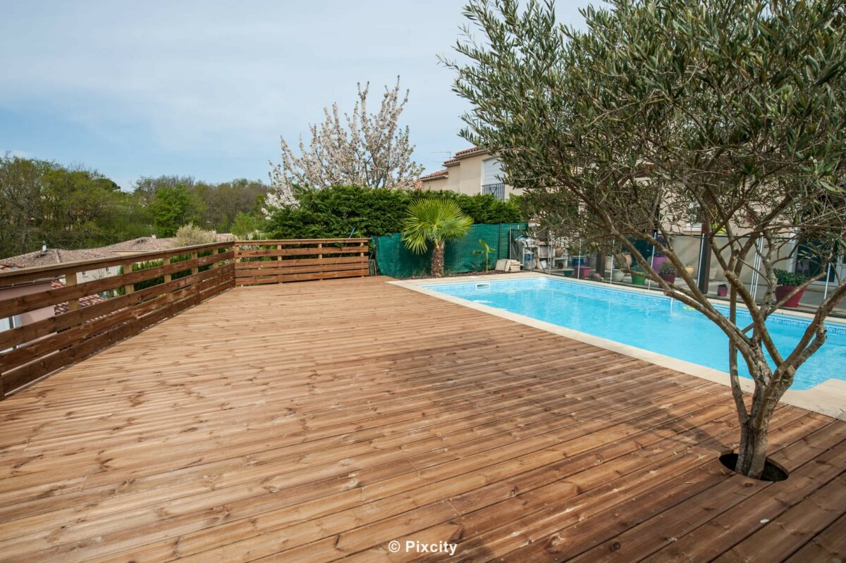 Aménagement terrasse bois autour piscine, grande terrasse bois pin naturel brut, olivier planté dans orifice terrasse, palissade brise-vue bois horizontal, garde-corps lattes bois horizontales, piscine rectangulaire bleue, pelouse, ciel gris doux, style naturel rustique