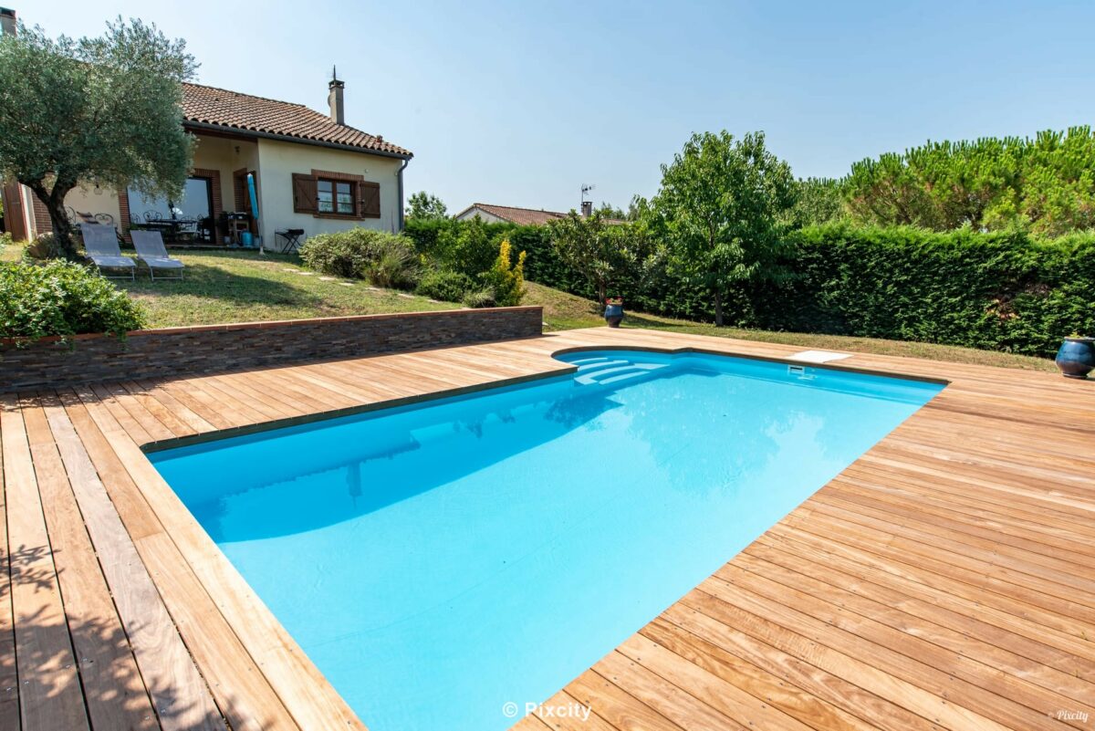 Aménagement terrasse bois autour piscine, gros plan piscine rectangulaire eau bleu profond cristallin, terrasse bois exotique teck large, haie cyprès denses taillés, pot céramique bleu marine artisanal, pelouse, ombre légère, ciel bleu, ambiance campagne