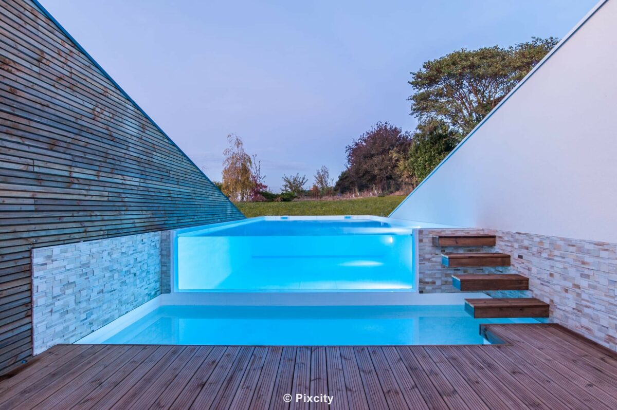 Aménagement terrasse bois autour piscine, piscine architecturale design paroi verre illuminée LED bleu électrique, façade bardage bois gris patiné, mur pierre parement clair, escalier flottant bois suspendu inox, terrasse bois, pelouse, ciel crépuscule bleu-violet, éclairage sous-marin nuit
