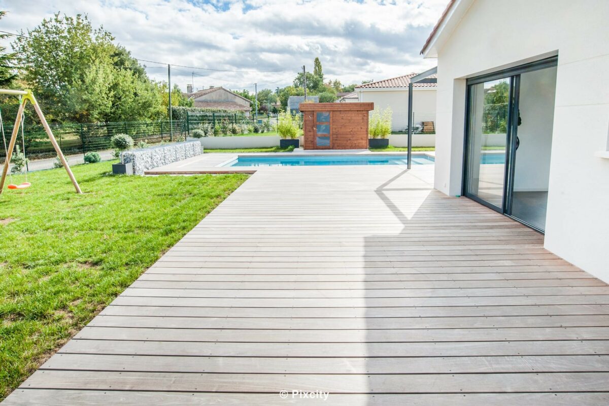 Aménagement terrasse bois autour piscine, grande terrasse composite gris ardoise spacieuse, piscine rectangulaire eau bleu clair en fond, maison contemporaine plain-pied enduit blanc, baie vitrée coulissante aluminium, pelouse verte, balançoire enfants bois, ciel nuageux dynamique