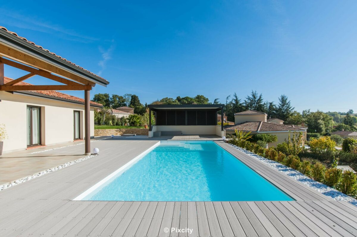 Aménagement terrasse bois autour piscine, grande piscine longue rectangulaire eau cristalline, terrasse composite gris clair, pergola bois massif poteaux ronds, pool house moderne baies vitrées noires, galets blancs, plantes ornementales, villa contemporaine, vue panoramique, ciel bleu