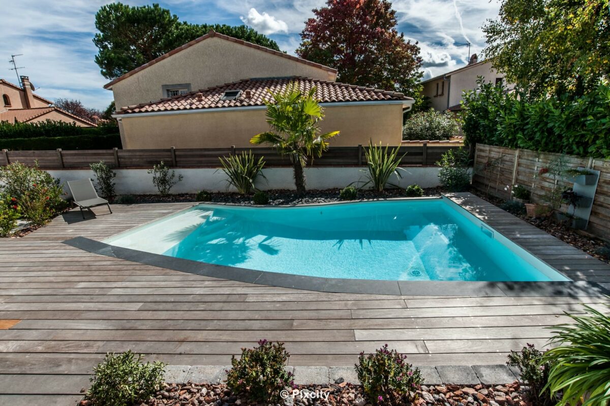 Aménagement terrasse bois autour piscine, piscine semi-ronde arrondie eau bleu-vert, terrasse composite gris ardoise, yucca palmier bananier plantes ornementales, galets bruns décoratifs, chaise longue noire design, maison résidentielle, ciel nuageux