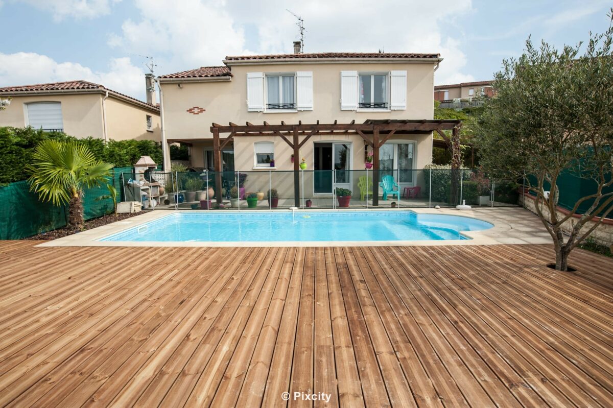 Aménagement terrasse bois autour piscine, grande terrasse bois pin traité autoclave, pergola bois poutres apparentes, piscine rectangulaire eau bleue, garde-corps verre transparent inox, oliviers méditerranéens, palmier, galets blancs, maison de ville, ciel bleu, printemps