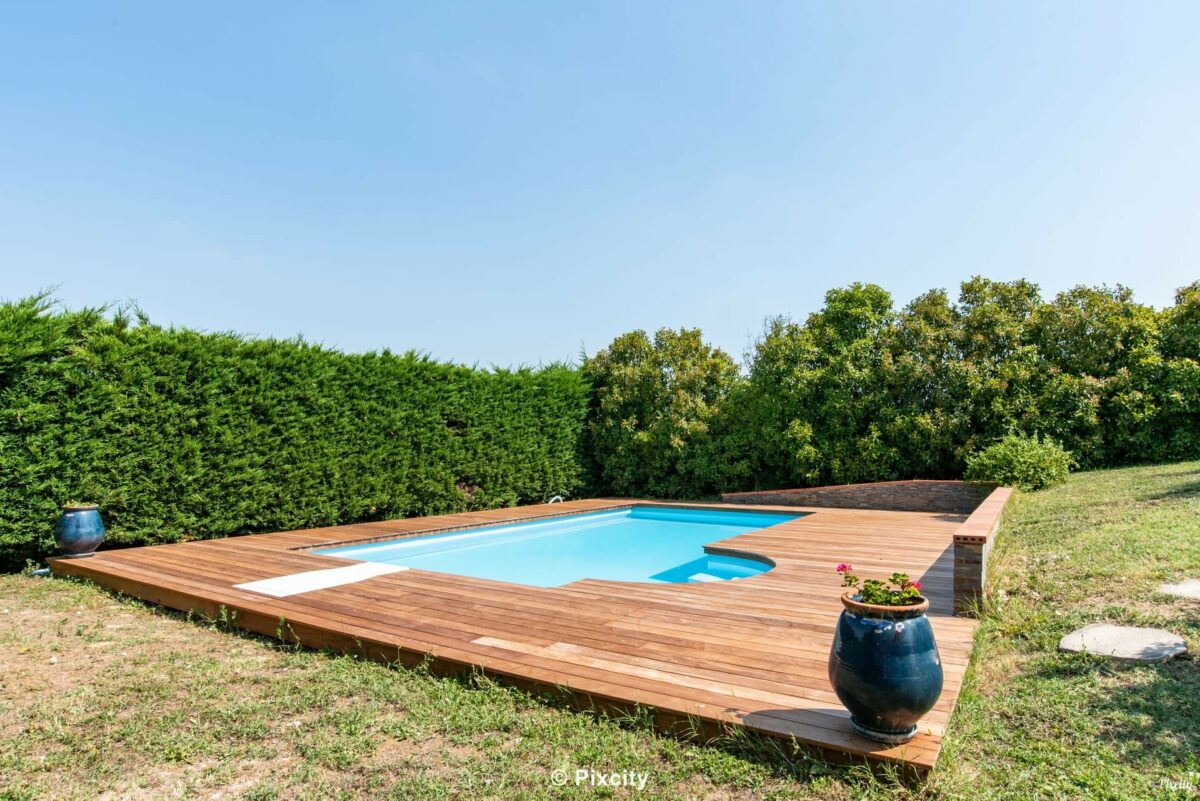 Aménagement terrasse bois autour piscine, piscine rectangulaire eau très bleue, terrasse bois exotique ipé clair, haie thuyas touffus bien taillés, pot céramique bleu pétrole artisanal, pelouse verte, ciel bleu immaculé, lumière plein soleil été campagne