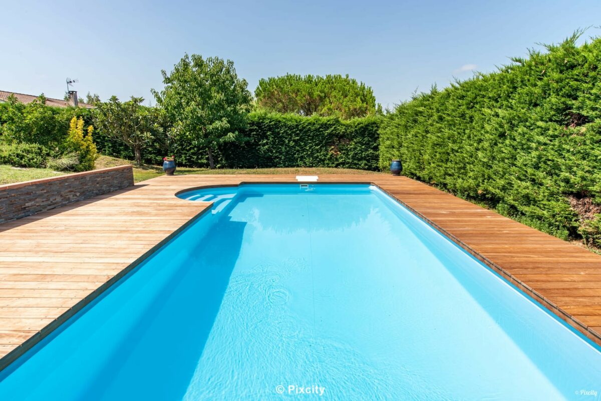 Aménagement terrasse bois autour piscine, piscine rectangulaire eau turquoise, grande terrasse bois exotique brun, haie haute thuyas taillés, galets blancs décoration bord piscine, pot céramique, ardoise bords, pelouse, ambiance mas gîte été