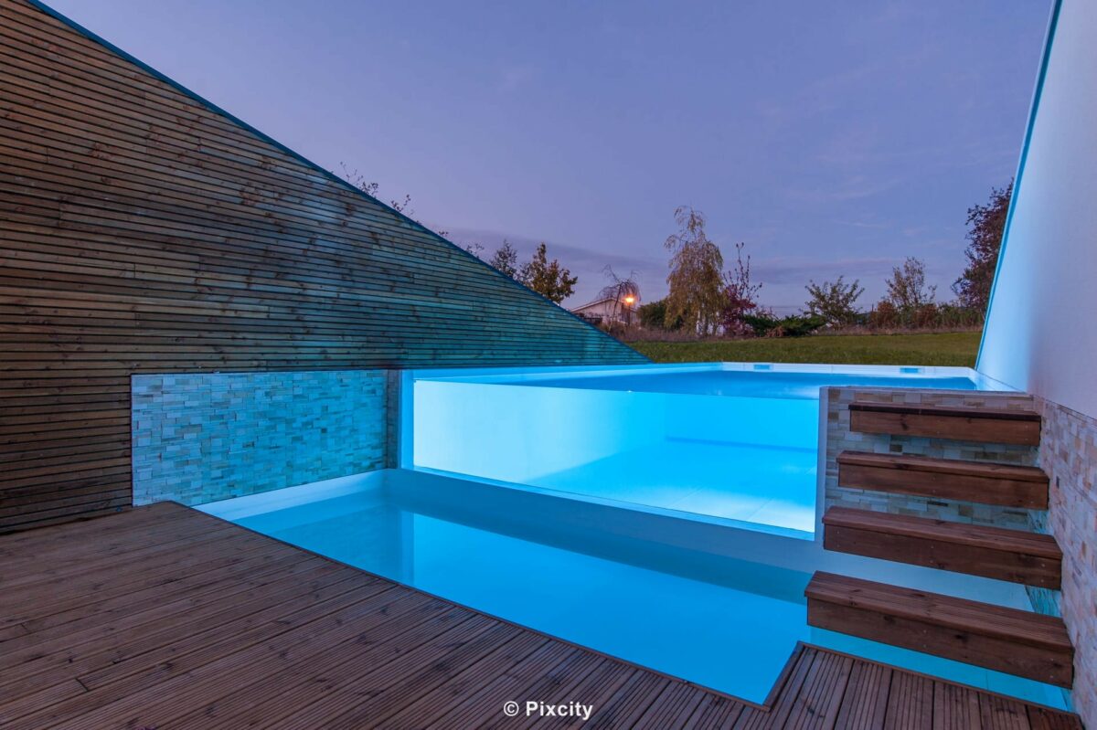 Aménagement terrasse bois autour piscine, piscine architecturale paroi verre illuminée bleue nuit, mur bardage bois horizontal patiné et pierre parement gris, escalier bois flottant suspendu marches, terrasse bois, ciel crépuscule mauve violet, éclairage LED submersible bleu électrique nuit