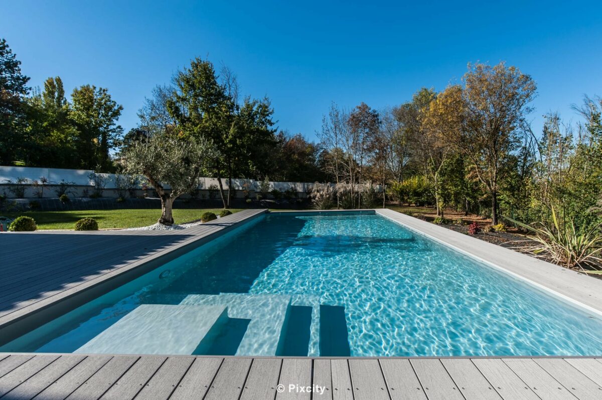 Aménagement terrasse bois autour piscine, très grande piscine rectangulaire longue eau cristalline, terrasse composite gris clair, villa contemporaine provençale toiture plate bardage bois brun, pool house fermé baies vitrées noires, galets blancs, plantes méditerranéennes, ciel bleu, vue collines