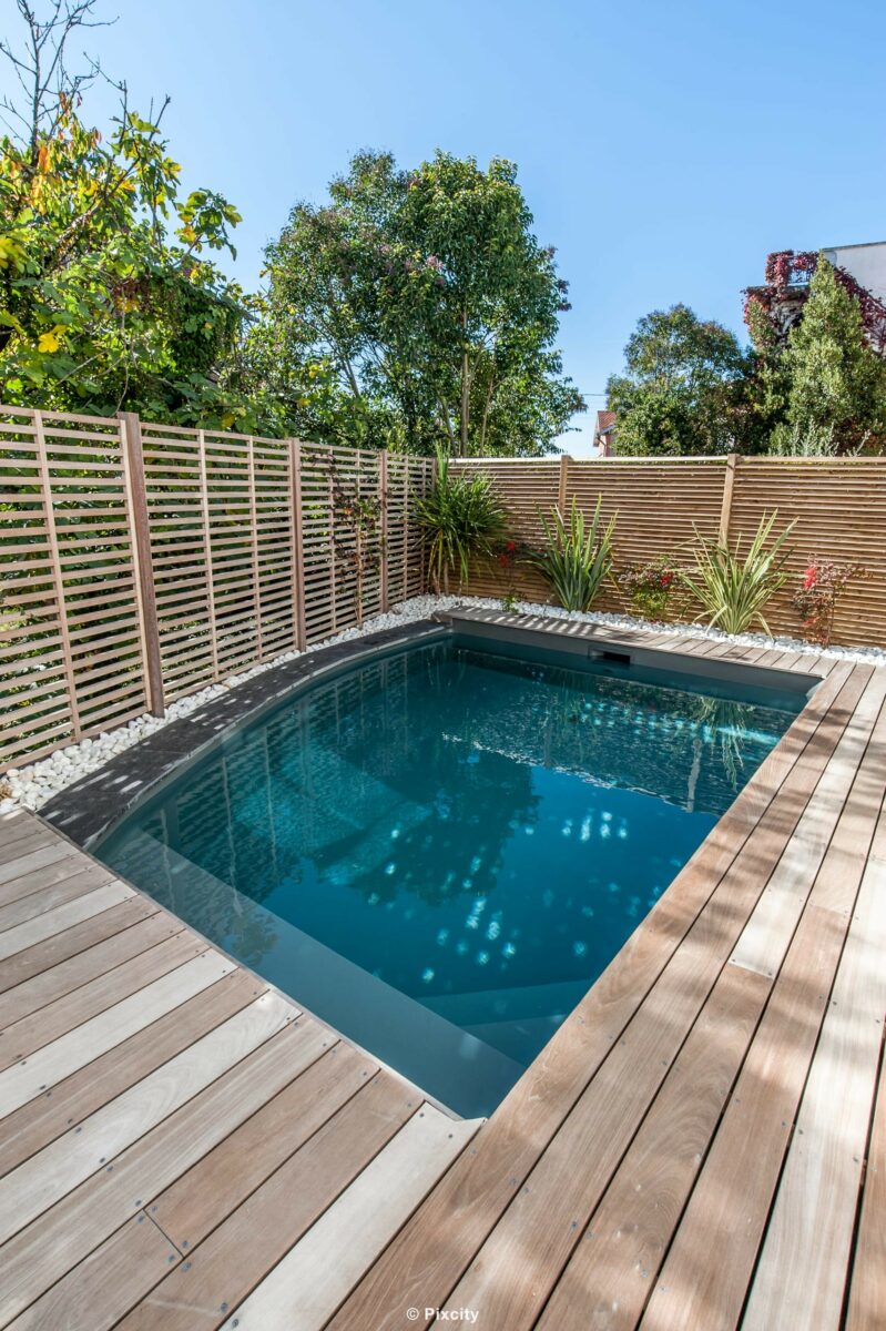 Aménagement terrasse bois autour piscine, petite piscine carrée fond gris eau bleu foncé, terrasse bois gris patiné, palissade brise-vue bois horizontal, dracaena yucca palmier exotiques en pot, galets décoratifs, maison blanche, pot rouge design, ciel bleu, jardinet citadin