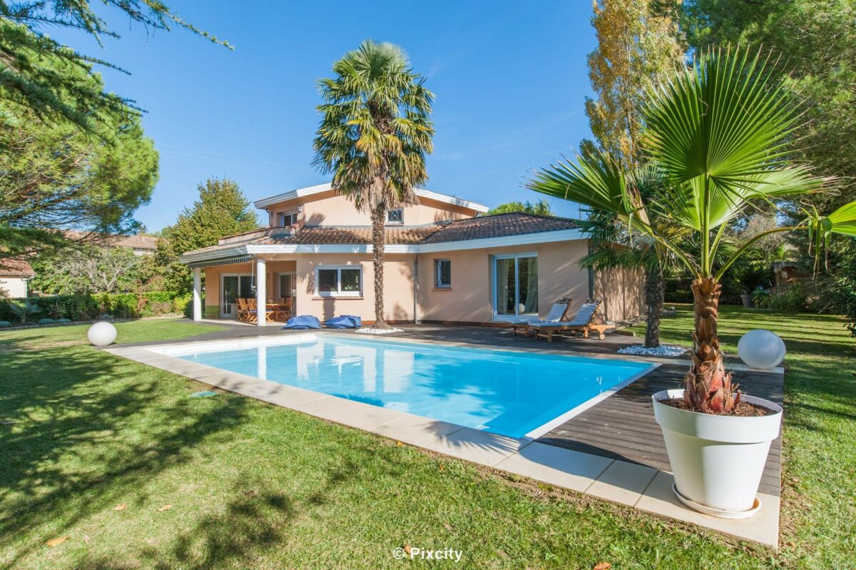 Aménagement terrasse bois autour piscine, villa résidentielle, piscine rectangulaire eau bleue, terrasse composite brun rouge, palmier trachycarpus, palmier washingtonia, pin parasol, chaises longues bleues design, pot blanc, galets, pelouse, jardin soigné