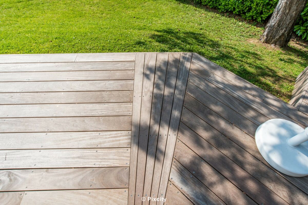 Aménagement terrasse bois autour piscine, vue aérienne plongée terrasse bois composite gris clair, lames croisées deux directions perpendiculaires, pelouse verte, table ronde blanche, ombres arbre projetées, lumière zénithale forte, géométrie épurée minimaliste
