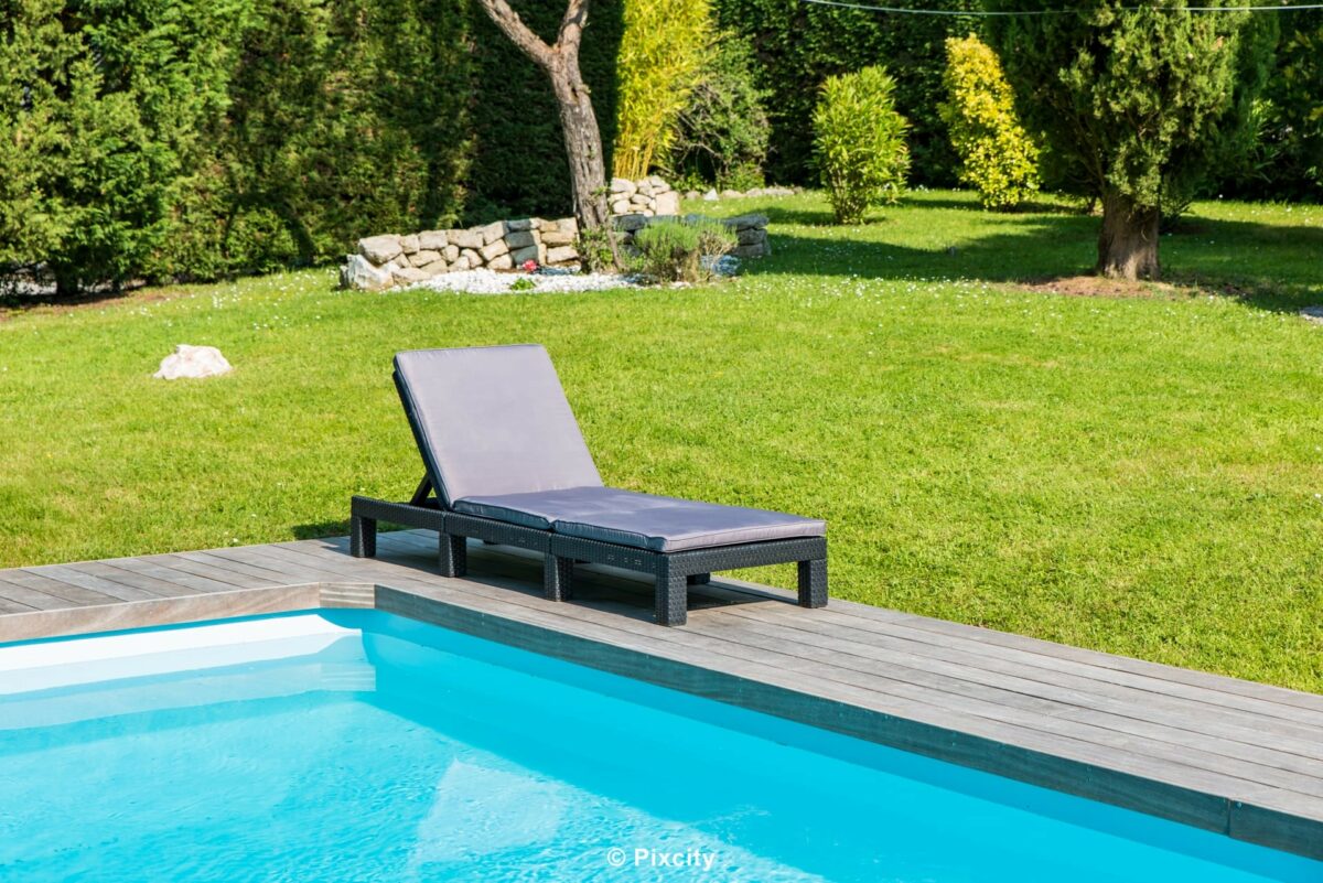 Aménagement terrasse bois autour piscine, chaise longue anthracite noire design bord piscine eau turquoise, terrasse bois gris naturel, pelouse verte bien entretenue, haie thuyas verts denses, arbres feuillus, lumière naturelle estivale, ombre légère